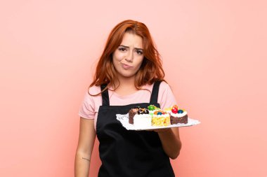 Genç redhead kız üzgün ifade ile izole pembe arka plan üzerinde farklı mini kek çok tutan