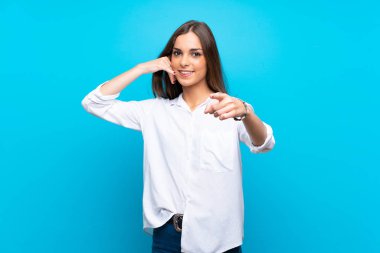 İzole edilmiş mavi arka planda genç bir kadın telefon hareketi yapıyor ve ön tarafı işaret ediyor.