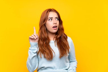Izole sarı arka plan üzerinde genç redhead kız bir fikir parmağını işaret düşünme