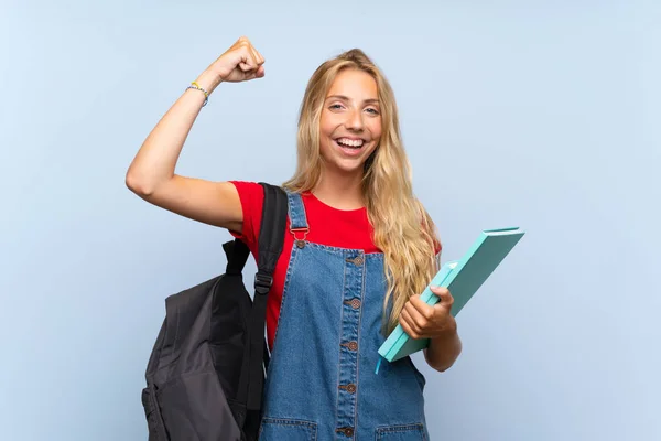 Young blonde student woman over isolated blue wall celebrating a victory