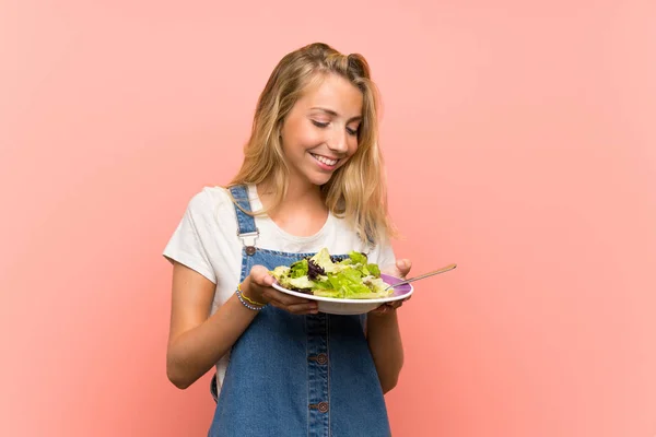 Gelukkige blonde jonge vrouw met salade over geïsoleerde roze muur — Stockfoto