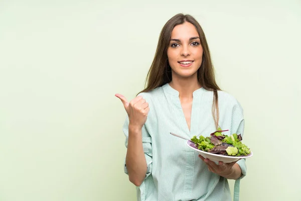 Young woman with salad over isolated green wall pointing to the side to present a product