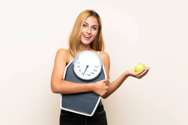 Jonge blonde vrouw met weegmachine en met een appel over geïsoleerde achtergrond — Stockfoto