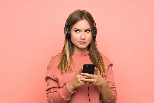Ragazza adolescente che ascolta musica con un cellulare su sfondo rosa isolato — Foto Stock