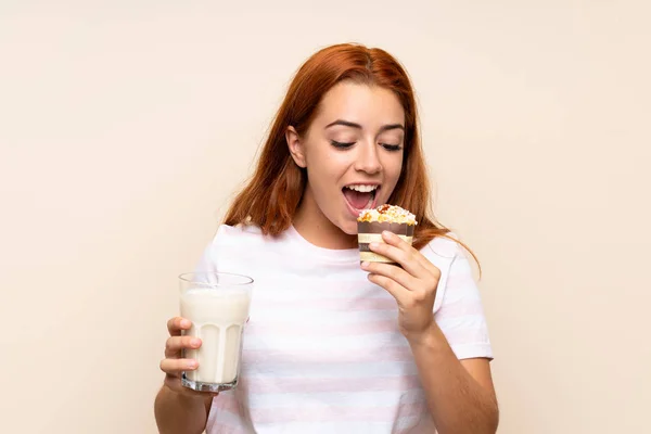 Adolescente pelirroja sosteniendo un vaso de leche y un panecillo sobre un fondo aislado — Foto de Stock