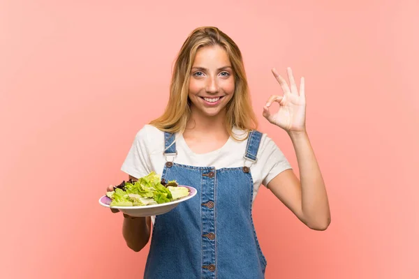 金发年轻女子与沙拉在孤立的粉红色墙壁显示与手指的确定标志 — 图库照片