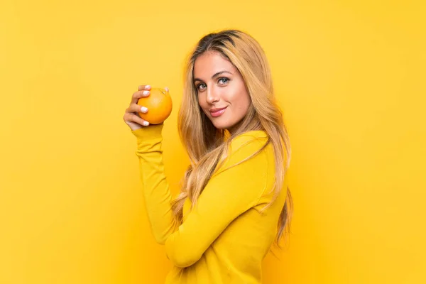 Junge blonde Frau hält eine Orange über gelbem Hintergrund — Stockfoto