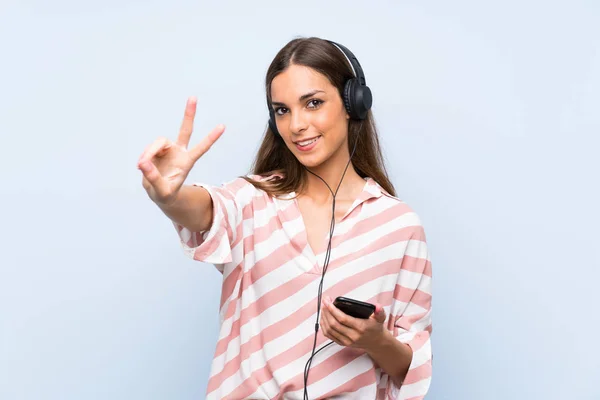 Young woman listening music with a mobile over isolated blue wall smiling and showing victory sign