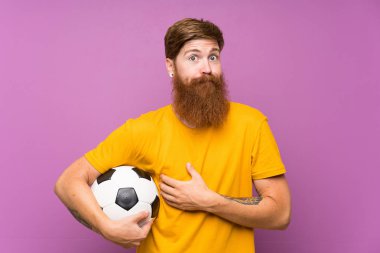 Uzun sakallı, futbol topunu izole edilmiş mor duvarın üzerinde tutan kızıl saçlı adam omuzlarını kaldırırken şüphe uyandırıyor.