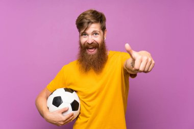 Kızıl saçlı, uzun sakallı bir adam futbol topunu izole edilmiş mor duvarın üzerinde tutuyor. Başparmakları havada, çünkü iyi bir şey oldu.