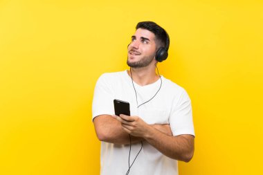 Genç yakışıklı adam gülümserken yukarı bakıyor izole sarı duvar üzerinde bir cep ile müzik dinliyor