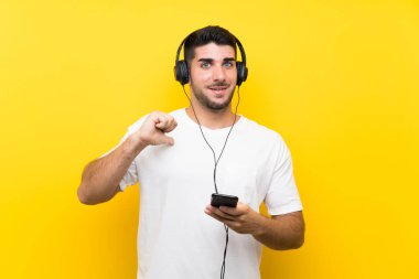 Genç yakışıklı adam izole sarı duvar üzerinde bir cep ile müzik dinleme gururlu ve kendini memnun