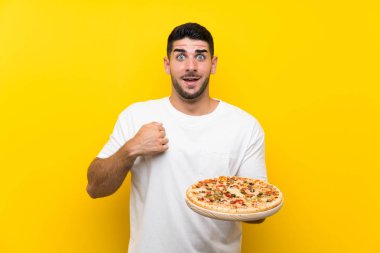 Genç yakışıklı adam sürpriz yüz ifadesi ile izole sarı duvar üzerinde bir pizza tutan
