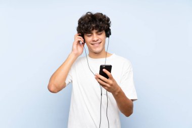 İzole mavi duvar üzerinde bir cep ile müzik dinleyen genç adam