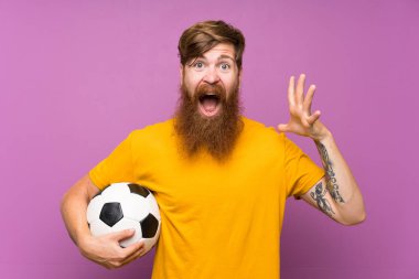 Kızıl saçlı, uzun sakallı, futbol topunu yüzünde şok ifadesiyle izole edilmiş mor duvarın üzerinde tutan adam.