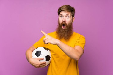 Uzun sakallı, uzun sakallı, futbol topunu izole edilmiş mor duvarın üzerinde tutan kızıl saçlı adam şaşırmış ve işaret ediyor.