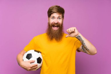 Kızıl saçlı, uzun sakallı, futbol topunu izole edilmiş mor duvarın üzerinde tutan gururlu ve kendini beğenmiş bir adam.