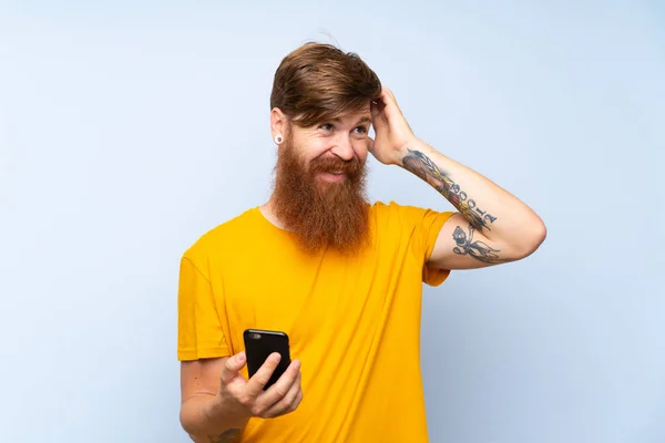 Redhead man with long beard with a mobile over isolated blue wall having doubts and with confuse face expression