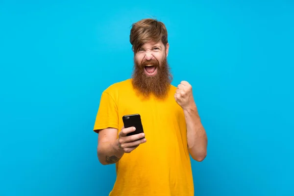 Ryšavý Muž Dlouhými Vousy Přes Izolované Modré Pozadí Telefonem Vítězné — Stock fotografie