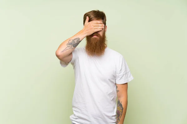 Redhead man with long beard over isolated green background covering eyes by hands. Do not want to see something