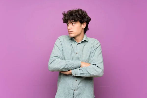 Young man over isolated purple wall making doubts gesture while lifting the shoulders