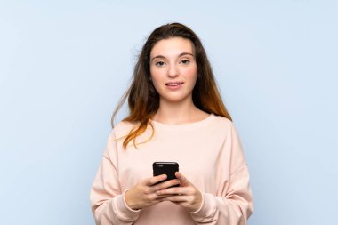 Genç esmer kız izole edilmiş mavi arka planda cep telefonuyla mesaj ya da e-posta gönderiyor.