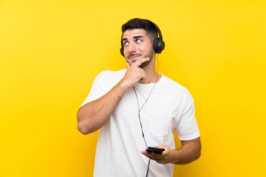 Genç yakışıklı adam izole sarı duvar üzerinde bir cep ile müzik dinleme bir fikir düşünme
