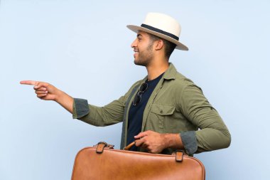 Genç yakışıklı adam izole mavi arka plan üzerinde bir ürün sunmak için yan işaret üzerinde bir vintage evrak çantası tutan