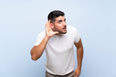 Kafkasyalı yakışıklı adam mavi arka planda elini kulağına koyarak bir şeyler dinliyor.