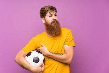 Kızıl saçlı, uzun sakallı, futbol topunu izole edilmiş mor duvarın üzerinde tutan adam gülümserken yukarı bakıyor.