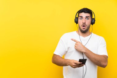 Genç yakışıklı adam izole sarı duvar üzerinde bir cep telefonu ile müzik dinlerken şaşırttı ve tarafı işaret