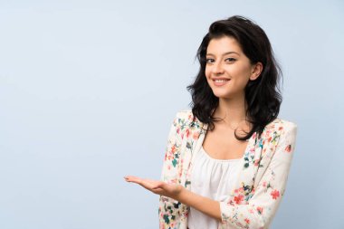 İzole edilmiş mavi arka planda genç bir kadın. Davet etmek için ellerini yana uzatıyor.