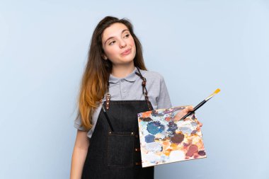 Genç esmer kız soyutlanmış mavi arka planda bir palet tutuyor ve bir fikir düşünüyor.