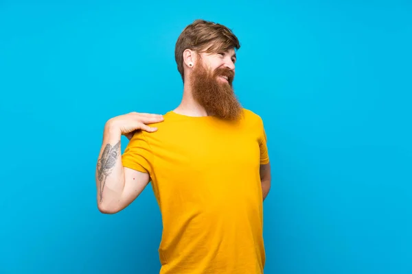 Redhead man with long beard over isolated blue background suffering from pain in shoulder for having made an effort
