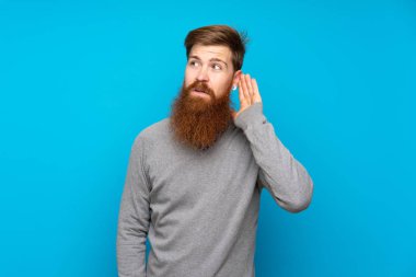 Uzun sakallı kızıl saçlı adam mavi arka planda elini kulağına koyarak bir şeyler dinliyor.