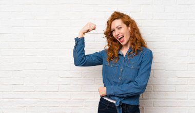 Kızıl saçlı kadın beyaz tuğla duvarın üzerinde güçlü bir jest yapıyor.