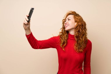 Boğazlı kazaklı kızıl saçlı kadın cep telefonuyla selfie yapıyor.