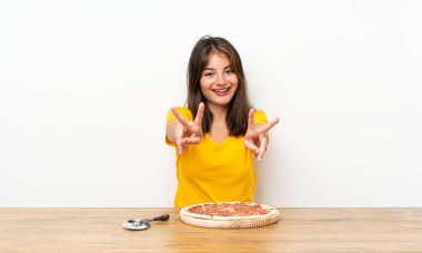 Elinde pizzayla gülümseyen ve zafer işareti gösteren beyaz bir kız.