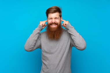 Uzun sakallı kızıl saçlı adam mavi arka planda hayal kırıklığına uğramış ve kulaklarını kapatmış.