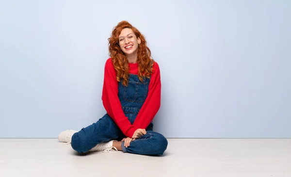 Redhead woman with overalls sitting on the floor with glasses and happy
