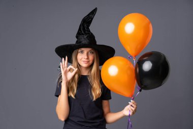 Young witch holding black and orange air balloons showing an ok sign with fingers