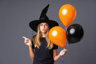 Elinde siyah ve turuncu hava balonları olan genç cadı yandaki kuşkuları işaret ediyor.