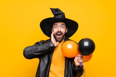 Cadı şapkalı bir adam cadılar bayramı partisi için elinde siyah ve turuncu hava balonları tutuyor. Şaşırmış ve şok olmuş bir yüz ifadesi var.
