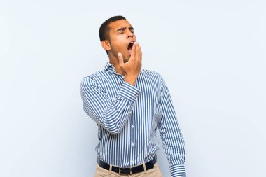 Genç, yakışıklı esmer adam, izole edilmiş mavi arka planda esniyor ve geniş ağzını eliyle kapatıyor.