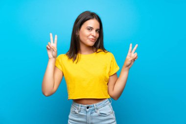 İzole edilmiş mavi arka planda genç bir kız zafer işaretini iki eliyle gösteriyor.