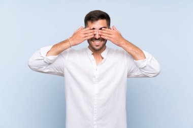 İzole edilmiş mavi arka planda sakallı yakışıklı bir adam. Gözlerini elleriyle kapatıyor.