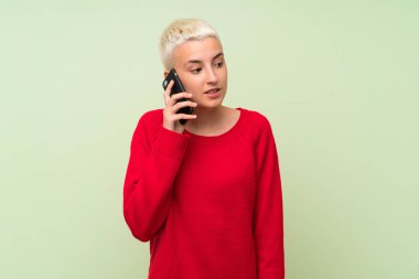 Yeşil duvarın üzerinde beyaz kısa saçlı genç kız cep telefonuyla sohbet ediyor.