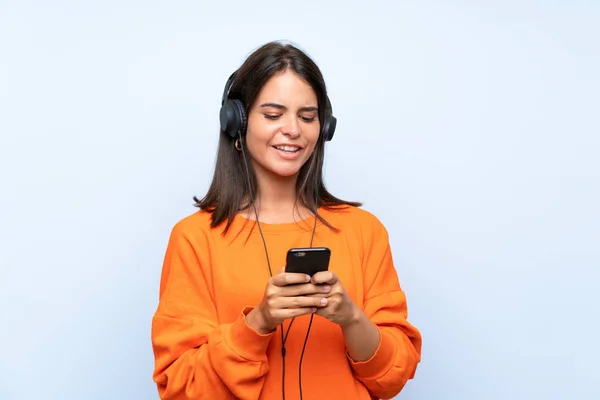 Mujer joven escuchando música con un móvil sobre una pared azul aislada —  Fotos de Stock