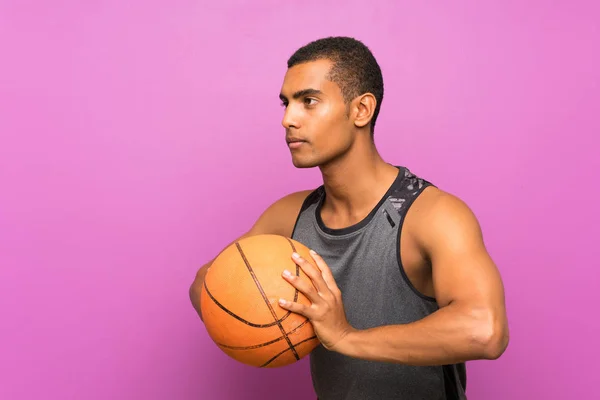 Pemuda olahraga dengan bola basket di atas dinding ungu terisolasi — Stok Foto