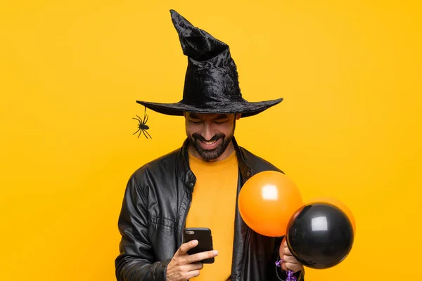Man with witch hat holding black and orange air balloons for halloween party sending a message with the mobile